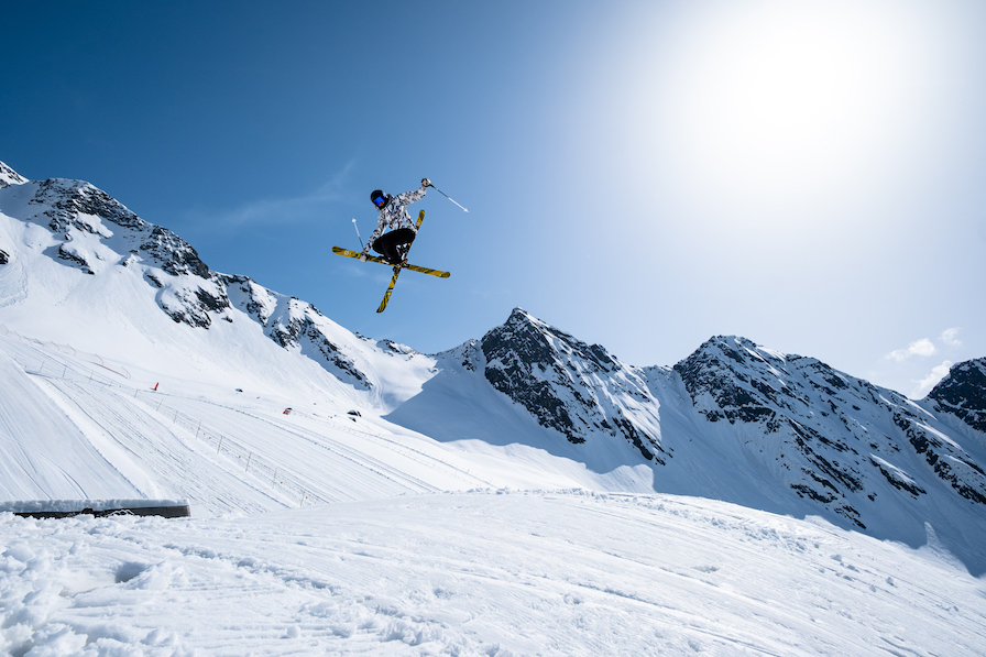 Skier sending a huge mute grab on a big jump with blue skies and a mountain range in the background